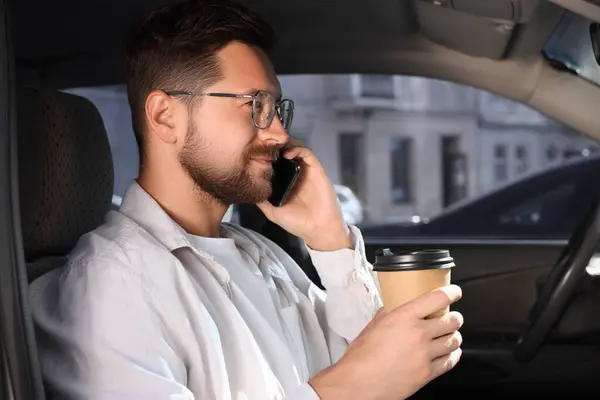 Coffee to go. Handsome man with paper cup of drink talking on smartphone in car