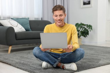 İçerideki halının üstünde bilgisayarla görüntülü konuşma yapan mutlu genç adam.