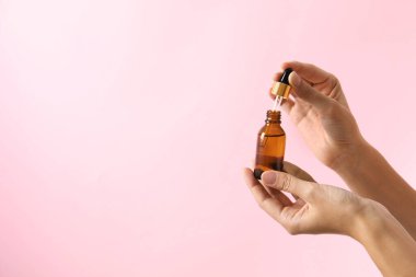 Pembe arka planda bir şişe kozmetik serumu ve damlatan kadın, yakın plan. Metin için boşluk