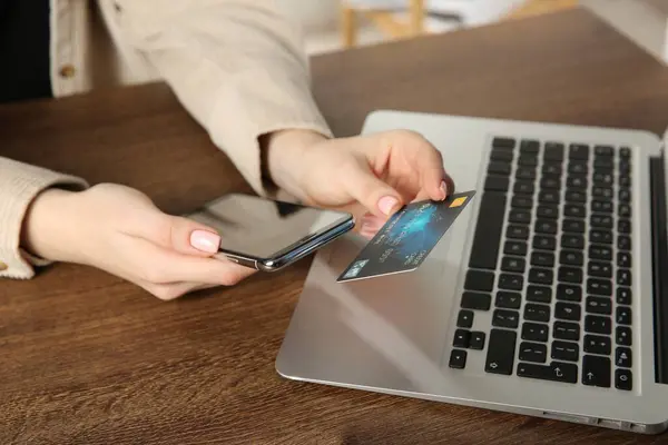 Online payment. Woman using smartphone and credit card near laptop at wooden table indoors, closeup