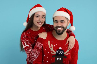 Mutlu genç çift Noel süveteri ve açık mavi arka planda Noel Baba şapkası giyer.