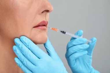 Doktor açık gri arka planda yaşlı kadına dudak iğnesi yapıyor, yakın plan. Estetik ameliyat