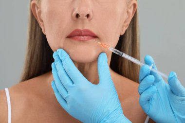 Doktor açık gri arka planda yaşlı kadına dudak iğnesi yapıyor, yakın plan. Estetik ameliyat