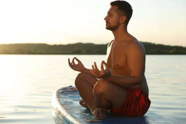 Adam gün batımında nehirde SUP tahtasında meditasyon yapıyor.