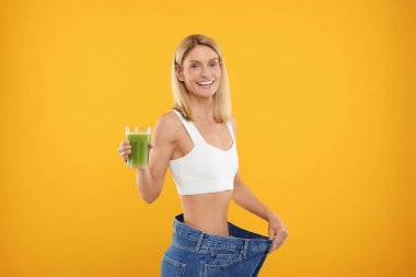 Bir bardak taze kereviz suyuyla ince bir kadın turuncu arka planda büyük kot pantolon giyiyor.