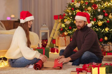 Noel Baba şapkalı mutlu çift evde ambalaj kağıdıyla Noel hediyesini süslüyor.