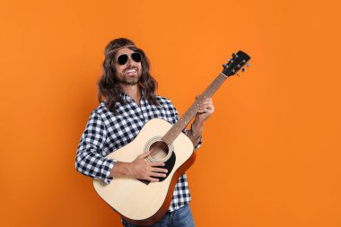 Güneş gözlüklü hippi adam turuncu arka planda gitar çalıyor.