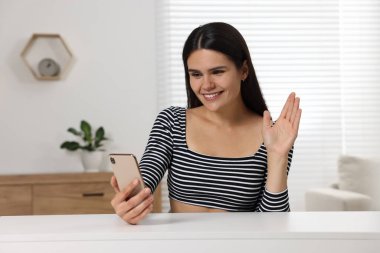 Akıllı telefondan görüntülü konuşma yapan ve odadaki masada el sallayan mutlu genç bir kadın.