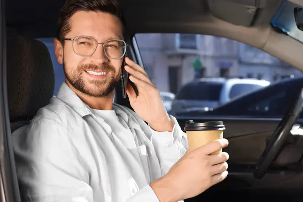 Coffee to go. Happy man with paper cup of drink talking on smartphone in car