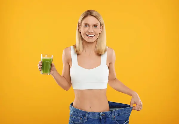Slim woman with glass of fresh celery juice wearing big jeans on orange background