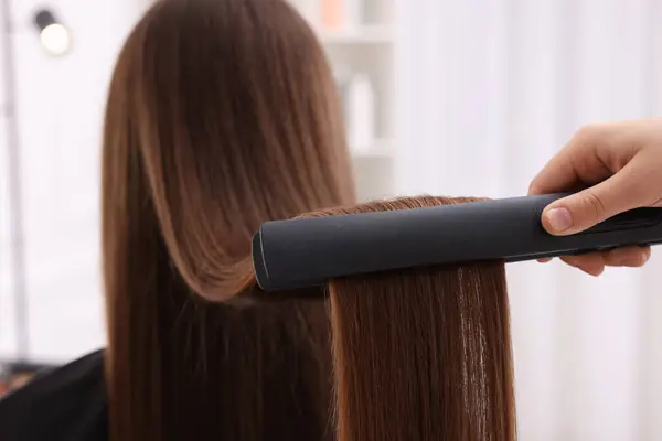 Hairdresser straightening woman\'s hair with flat iron indoors, closeup