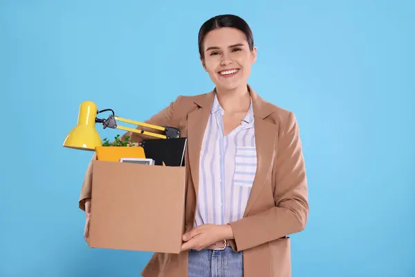 Happy unemployed woman with box of personal office belongings on light blue background