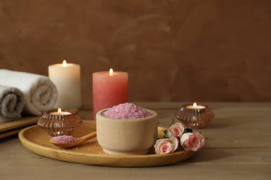 Bir kase pembe deniz tuzu, güller, yanan mumlar, bitkisel masaj torbaları ve ahşap masadaki havlular.