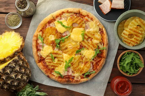 Delicious pineapple pizza and ingredients on wooden table, flat lay