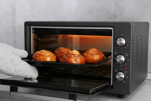 Woman taking out baking pan with delicious spiral buns from electric oven, closeup