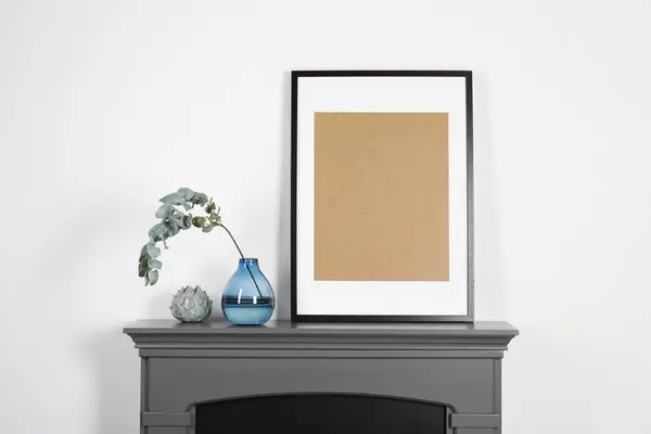 Empty frame and eucalyptus branch in vase on fireplace near white wall indoors. Interior design
