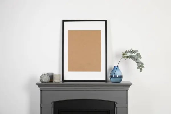 Empty frame, candles and eucalyptus branch in vase on fireplace near white wall indoors. Interior design