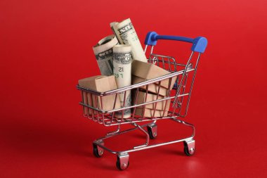 Kırmızı arka planda karton kutular ve dolar banknotları olan küçük metal alışveriş arabası.