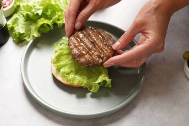 Açık gri masada kızarmış köfte, marul ve çörekle lezzetli hamburger yapan kadın, yakın plan.