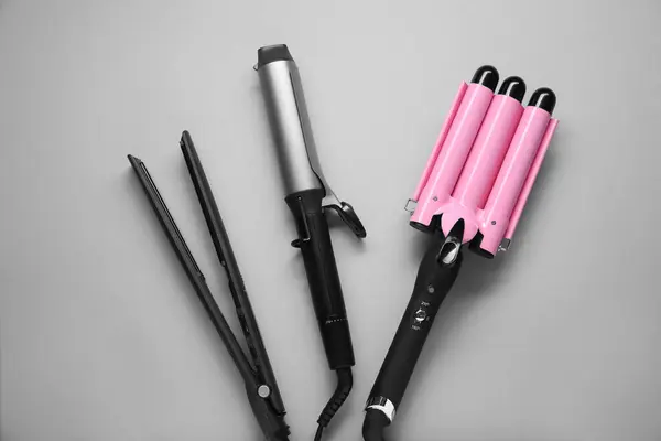 Different curling irons and hair straightener on grey background, flat lay