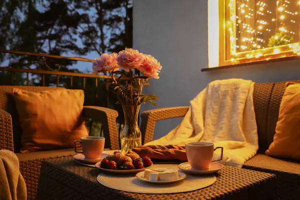 Rattan table with drink, food and flowers on outdoor terrace in evening
