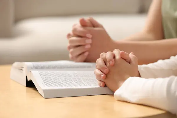Girl and her godparent praying over Bible together at table indoors, closeup. Space for text