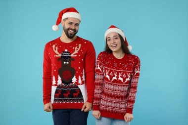 Noel Baba şapkalı mutlu çift açık mavi arka planda Noel süveterlerini gösteriyor.