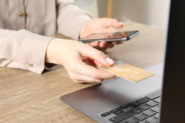 Çevrimiçi ödeme. Akıllı telefon ve kredi kartı kullanan bir kadın laptopun yanında kapalı masa başında, yakın plan.