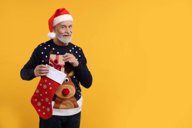 Noel süveteri ve Noel Baba şapkası giyen son sınıf öğrencisi turuncu arka plandaki çoraptan hediye alıyor. Metin için boşluk