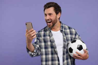 Futbol topu ve mor arka planda akıllı telefonu olan duygusal spor fanatiği.