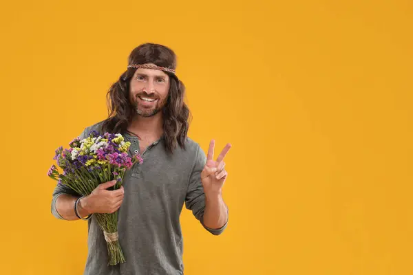 Hippie man with bouquet of colorful flowers showing V-sign on orange background, space for text