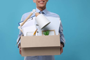Açık mavi arka planda kişisel ofis eşyalarıyla dolu bir kutuya sahip işsiz genç adam, yakın plan.