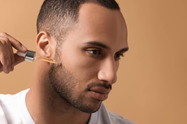 Açık kahverengi arka planda yüzüne kozmetik serumu süren yakışıklı bir adam. Yakın plan. Metin için boşluk