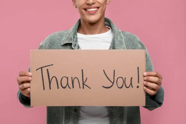 Man holding cardboard sheet with phrase Thank You on pink background, closeup