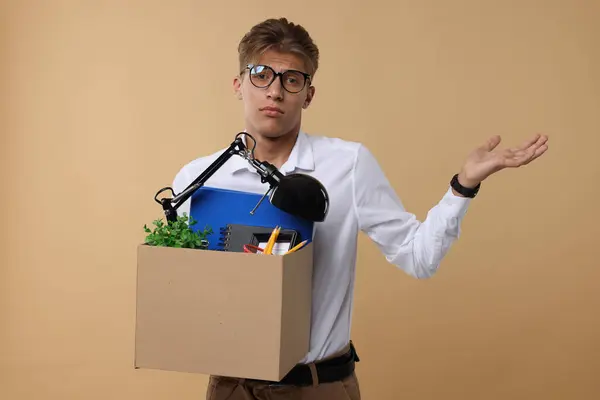 Confused unemployed young man with box of personal office belongings on beige background