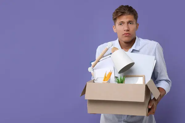 Unemployed young man with box of personal office belongings on purple background. Space for text