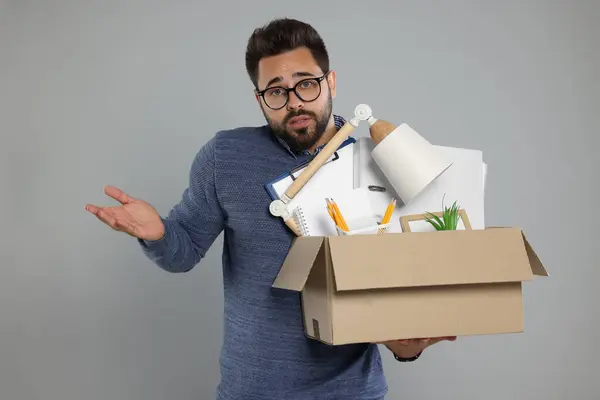 Confused unemployed man with box of personal office belongings on light grey background