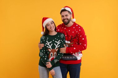 Noel süveteri ve turuncu arka planda Noel Baba şapkası giyen mutlu çift.