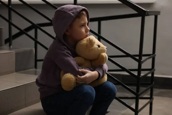 Stock image Child abuse. Upset boy with teddy bear sitting on stairs indoors