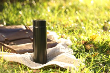 Black thermos and bag with blanket on green grass outdoors, space for text clipart