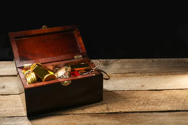 Treasure chest with gold bars, coins, jewelry and gemstones on wooden table. Space for text