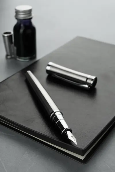 Stylish fountain pen, bottle of ink, cap and leather notebook on light grey textured table