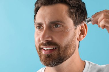 Gülümseyen adam açık mavi arka planda yüzüne kozmetik serumu uyguluyor.