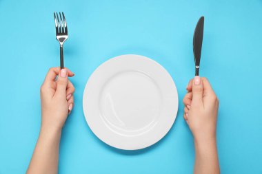 Çatal ve bıçak taşıyan bir kadın açık mavi masada boş tabağın yanında, üst tarafta.