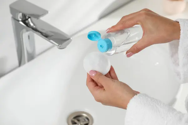 Woman pouring makeup remover from bottle onto cotton pad near sink indoors, closeup
