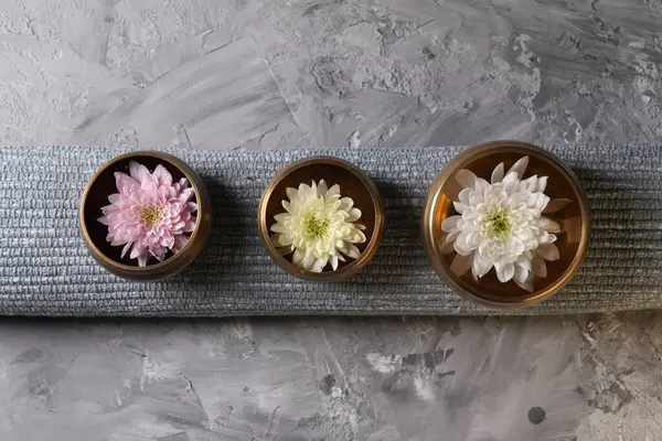 Tibetan singing bowls with water, beautiful chrysanthemum flowers on grey textured table, top view