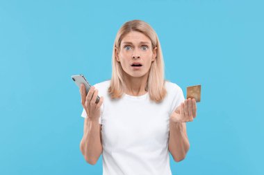 Açık mavi arka planda kredi kartı ve akıllı telefonu olan stresli bir kadın. Dikkatli ol, sahtekar.