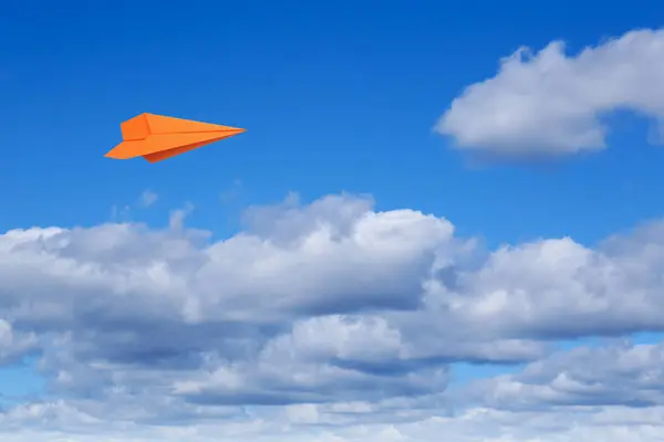 Orange paper plane flying in blue sky with clouds