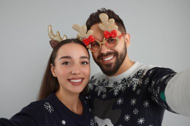 Mutlu genç çift, Noel süveteri, ren geyiği saç bandı ve gri arka planda selfie çeken komik gözlükler.