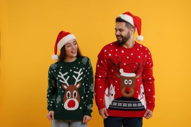Noel Baba şapkalı mutlu çift turuncu arka planda Noel süveteri gösteriyor.